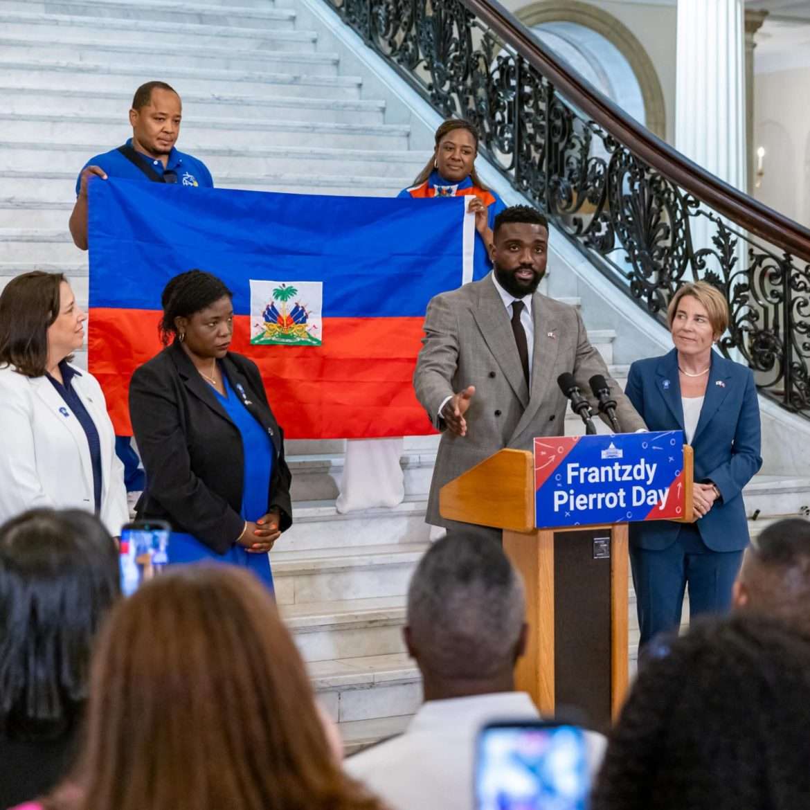 Frantzdy Pierrot s'adresse au public lors de la cérémonie de proclamation du « Frantzdy Pierrot Day » au State House de Boston, devant le drapeau haïtien, entouré de la gouverneure Maura Healey et de personnalités de la diaspora. © DR